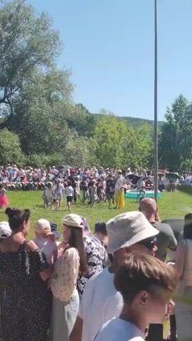 This video captures an outdoor event with a large crowd of people gathered on a grassy field under a clear blue sky. In the foreground, several individuals are partially visible, facing away from the camera. They appear to be spectators. Further in the background, a group of children and adults are actively participating in what looks like a performance or game. One individual in the center, wearing a shiny, possibly sequined outfit and a yellow hat, is standing and gesturing, suggesting they are the focal point of the activity. Other adults, some in uniforms or distinguishing clothing, are also present with the children, possibly as organizers or supervisors. The environment is characterized by lush green grass, trees in the background, and a gently sloping hill. The overall atmosphere appears festive and communal.