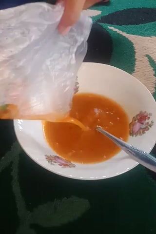 A hand is pouring the contents of a clear plastic bag into a white bowl. The bowl contains orange broth and a metal spoon. The contents of the bag appear to be noodles, vegetables, and meat. The bowl is sitting on a green patterned surface.