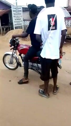 The video shows a street scene in what appears to be a small town.  A man in a red shirt sits on a motorcycle talking to another man.  The camera pans across the street showing a building with a purple roof, then a view of a tower and a building that says "Oluin Palace". There are several people visible in the shots. The video is shot on a cloudy day on a dirt road.