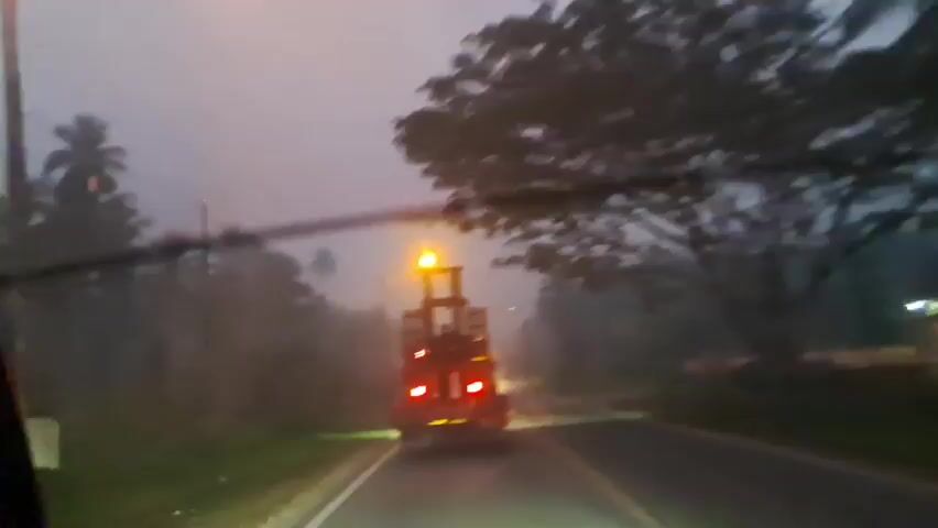 The video shows a view from inside a vehicle, looking forward through a windshield. A large, red forklift is traveling ahead on a two-lane road. The forklift has its brake lights illuminated and is facing away from the camera. The scene appears to be at dusk or dawn, with low visibility due to fog or mist. The road is wet, suggesting it may be raining. Tall trees line both sides of the road, and there are power lines visible overhead. The overall atmosphere is dim and somewhat hazy.