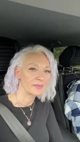 A woman with short, wavy, silver-purple hair and light makeup, wearing a black long-sleeved shirt and multiple necklaces, sits in the driver's seat of a car. She is looking directly at the camera and speaking. Her left hand is raised and gesturing with a pointed finger. The car's interior is visible, including the black leather headrest of the seat behind her and a gray seatbelt. A blue and white checkered fabric is visible on the passenger side. The background outside the car appears to be a blurred landscape with trees and sky.