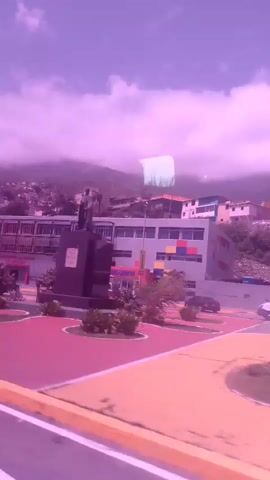 This video shows a view from a moving vehicle, likely a bus, passing through a town or city. The scene is characterized by buildings, some of which appear to be older and in disrepair, situated on a hillside. The sky is cloudy with a pinkish hue, suggesting it might be sunrise or sunset, or perhaps a filter has been applied to the footage.

In the foreground, there's a colorful, patterned area that could be a public plaza or park with geometric shapes. Several vehicles are visible, including white vans and cars, as well as what appears to be a truck. A statue of a person stands in a fountain area. The architecture of the buildings, with their various colors and styles, and the overall hillside setting are distinctive.

There are no easily discernible people in clear focus, though there might be figures in the distance or within the buildings. The activity is simply the vehicle's transit through the area. The prominent colors in the sky and the foreground pavement are notable. The location context of La Guaira, Venezuela, suggests a coastal or near-coastal town with a mountainous backdrop. The overall mood is somewhat subdued, possibly due to the lighting and the state of some of the structures. No legible text is visible.