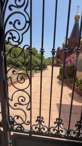 A person is visible standing near a horse in a garden with a church in the background. The camera is looking through a black wrought iron gate. The path is paved with light brown tiles. There are trees and bushes. The sky is blue and there are some clouds.