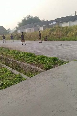 The video shows a group of people on an asphalt surface, possibly a road or court, with a strip of green vegetation on the right. Several individuals, appearing to be young males, are engaged in some activity. One person in a black shirt and shorts is standing near the center. Further down the path, another person in a black shirt and shorts is observed. Closer to the foreground, two individuals in dark shirts and shorts are visible, with one appearing to be in motion. In the background, there are houses with white walls and dark roofs, situated next to tall, dry grass. The lighting suggests it is daytime, possibly early morning or late afternoon, with an overcast sky. The perspective is angled, making it appear as if the camera is tilted.