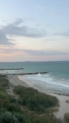 The video pans over a serene beach scene. The ocean is a vibrant turquoise, with gentle waves lapping the sandy shore. Several stone breakwaters jut out into the water, creating calm pockets along the coastline. Lush green bushes line the edge of the beach, adding a touch of natural beauty. As the camera continues to pan, rows of empty lounge chairs and umbrellas are revealed, suggesting a resort or a public beach preparing for visitors. The sky is a soft blend of pale blue and pink, with wispy clouds adding to the peaceful atmosphere. There are no people visible in the frame.