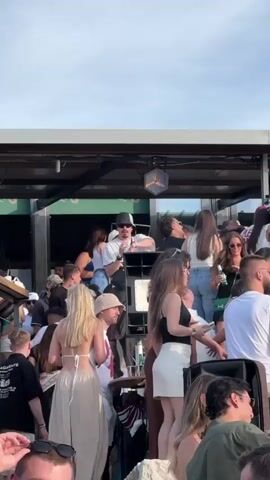 A crowd of young adults are gathered outdoors on a sunny day. They are standing on what appears to be a raised platform or rooftop, possibly at a party or event. In the background, a DJ is visible on a platform, wearing a white t-shirt and a black baseball cap, and is gesturing with his hands. He is standing next to a speaker system. The crowd is densely packed, with many people dressed in casual summer clothing. Some individuals are holding drinks and interacting with each other, suggesting a social gathering. The overall atmosphere appears lively and festive.