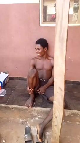 Two shirtless young men are sitting on a concrete step outside a building. The men are both dark-skinned. They are wearing dark shorts and sandals. One man is looking at a mobile phone in his hand. The other man is looking at the mobile phone. There is a red plastic cup next to one of the men. There is a white box and a plastic bottle on the ground next to the men. The wall behind them is a light reddish color.