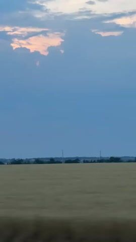 The video shows a wide-angle view of a landscape at dusk. The sky is a deep blue with scattered clouds that are tinged with pink and orange, indicating the setting sun. Below the sky, a flat expanse of dry, golden-brown grass or crops stretches across the frame, suggesting a rural or agricultural setting. A dark, thin line of trees or vegetation marks the horizon, separating the field from the sky. The image is slightly blurred, giving a sense of motion, possibly from being filmed from a moving vehicle. There are no people or distinct objects visible in the frame, just the natural elements of the sky, land, and distant vegetation.