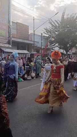 A group of young women in traditional Indonesian attire, likely from a parade or festival, are walking down a street. The woman in the foreground wears a vibrant orange headscarf, an ornate red and gold top, and a patterned skirt. She is followed by other women in similar elaborate costumes, many of whom are holding and displaying decorative fabric. They are walking on a paved road, with spectators seated on the side and buildings visible in the background. The sky is overcast with some sunlight breaking through. The overall atmosphere suggests a cultural celebration.