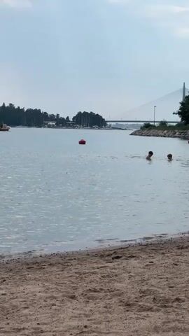 A sandy beach shore is in the foreground. The water is calm with some ripples. A red buoy floats in the water. Two people are swimming in the water. A boat is moving across the water in the distance. Trees line the shore in the distance. A bridge with a tall tower is visible in the background. The sky is overcast.