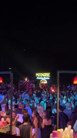 The video shows a large crowd of people at an outdoor party at night. A neon sign in the background reads "MOONRISE." The party appears lively with many people dancing and socializing under colored lights, including purple, green, and red. In the foreground, a bar is visible with drinks and a bartender standing behind it. Corona beer boxes are also visible near the bar. The overall setting suggests a vibrant nighttime social gathering with music and drinks.