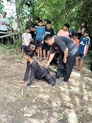 The video shows a group of people gathered in a wooded area, watching a man on his hands and knees. Another man is attending to him, apparently tying something to his ankle. The group watching is mostly young men and boys, with a few women visible in the background. The location appears to be a natural setting with trees, bamboo, and dirt ground. The man on the ground is wearing dark clothing and a headscarf, while the other man is dressed in a dark shirt and pants. There are also a few objects that look like knives or machetes on the ground near the men. The atmosphere seems curious, as the onlookers are focused on the activity taking place.