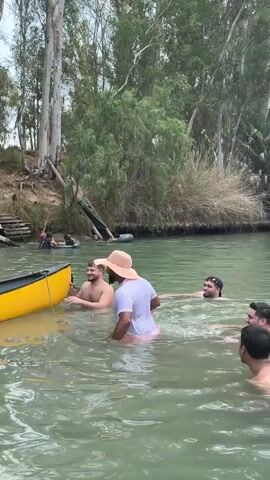 A group of people are in a river. One person is holding onto a yellow canoe, while another person in a white shirt and a hat is in the water next to them. There are other people in the river, some of whom are partially submerged. The river is surrounded by trees and lush vegetation. There are wooden stairs leading up to a bank on the left side of the image.