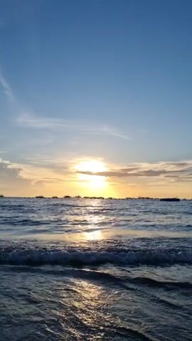 This media file captures a serene sunset over the ocean in Pattaya City, Thailand. The scene is dominated by the vast expanse of the sky and sea, with the sun positioned low on the horizon, casting a warm, golden glow. Wisps of clouds are scattered across the sky, adding texture to the brilliant blue canvas. The ocean is calm, with gentle waves lapping at the shore. The sunlight reflects off the water's surface, creating a shimmering path that leads towards the setting sun. In the distance, a line of boats and possibly other vessels are silhouetted against the vibrant sky, suggesting the presence of a busy harbor or coastline. The overall mood is peaceful and tranquil, evoking a sense of calm and natural beauty. The time of day is clearly sunset, indicated by the low position of the sun and the warm, diffused light. The location cues, such as the boats and the general seascape, are consistent with a coastal city like Pattaya. There is no visible text in this media file.