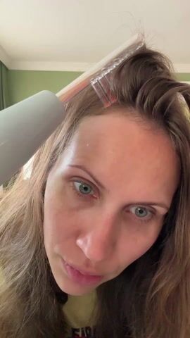 A woman is shown from the forehead up, using a device with a comb-like attachment on her scalp. The device emits a light and a buzzing sound, and she moves the comb through sections of her hair while looking directly at the camera with a slightly furrowed brow. The device is white and cylindrical, and the comb attachment is clear with an orange light visible within. The woman has fair skin, light brown hair, and light blue eyes. A green wall and a white ceiling are visible in the background.