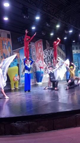 A group of performers are on a stage during a performance. There are two performers in red costumes, one in a yellow costume, one in a white dress, one in a black and white outfit, one in a blue dress, a character in a mouse costume, a character in a yellow clown costume, and one performer with white wings. The stage has a backdrop of buildings with signs that say "BROADWAY" and "JERSEY BOYS". There are stage lights above the performers. The performers are in various poses, some are lying down, and some are standing. The video is vertically oriented.