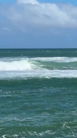 A video shows a wide shot of the ocean with moderate waves. The water is a deep greenish-blue color, and white foamy crests are visible as the waves break and roll towards the viewer. The horizon line is visible in the distance, separating the ocean from the sky. The sky is a clear blue with scattered white cumulus clouds. There is a faint, distant silhouette on the horizon, possibly land or a large vessel, but it is indistinct. The overall scene depicts a sunny day at sea. No people or specific objects are identifiable in the foreground or mid-ground. The waves are in constant motion, crashing and receding.