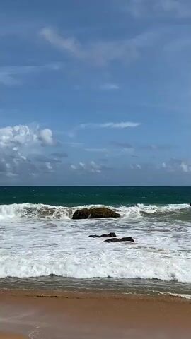 This media file captures a serene beach scene in Hikkaduwa, Sri Lanka. The foreground features the sandy shore, gently meeting the foamy white waves of the ocean. A few dark rocks are scattered in the shallow water, partially submerged as the waves crash and recede. In the mid-ground, larger rock formations are visible, breaking the turquoise water as waves roll in. The ocean stretches out to the horizon, meeting a sky filled with scattered white clouds against a backdrop of clear blue. The overall impression is one of a peaceful, natural environment, likely during daylight hours, given the bright sky. There are no people or man-made objects visible, focusing entirely on the natural beauty of the coastline. The movement of the waves and the texture of the foam suggest a dynamic yet tranquil atmosphere.