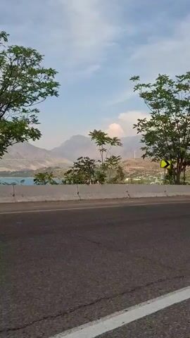 A side view from a moving vehicle on a road shows trees and a concrete barrier. The road has white lines marking the lanes. Beyond the barrier, there are more trees and a landscape with hills and a body of water. The sky is visible with some clouds. A yellow sign with a black arrow pointing right is visible on a post near the trees. The video captures the scenery as the vehicle moves forward.