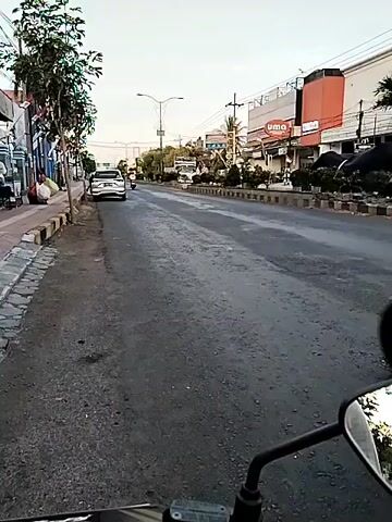 The video is a first-person perspective from a motorcycle, looking forward down a street with shops and buildings on the right. The road is paved and appears to be slightly wet. There are several vehicles on the road: a white car in the distance, a scooter with a rider in a yellow shirt in the middle of the frame, and a white truck with a box trailer passing by. A yellow vehicle is also visible briefly on the right side of the road. The sky is overcast. The rider's left hand and part of the motorcycle's handlebar and mirror are visible in the foreground. The mirror reflects a view of the road and traffic behind.