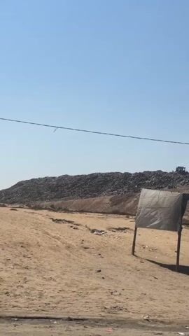 The video shows a wide-angle view of a landfill under a clear blue sky. In the foreground, there is a dusty, uneven terrain with scattered debris. To the right, a makeshift shelter with a canvas roof is set up, appearing to be a small stall or shop with various items displayed. The shelter is constructed with poles and fabric. In the middle ground, a large mound of garbage is visible, with a bulldozer working on top of it. The terrain leading up to the garbage mound is also dusty and appears to be a dirt embankment. The overall impression is one of an industrial or waste disposal area in a dry, arid environment.