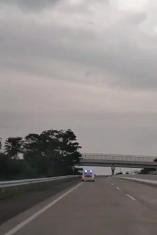 A white car with a blue light bar on its roof drives away from the camera on a multi-lane highway under an overcast sky. The vehicle is seen driving towards and then under an overpass. Trees line the left side of the road, and a guardrail is also visible. A yellow, diamond-shaped road sign is partially obscured on the left, and a blue rectangular sign is visible under the overpass but unreadable. The video captures the car's movement from behind as it progresses along the road.