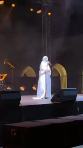 A woman wearing a white outfit and hijab is performing on stage. She is holding a microphone and singing. The stage is lit with spotlights. There is a band set up to her left and speakers in front of the stage. Behind her is a backdrop of arches lit in gold. There is a metal tower structure above the stage. A person in black walks in front of the camera momentarily.