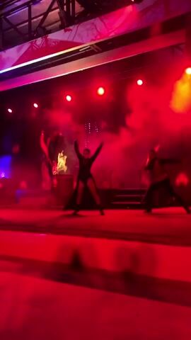 The video captures a performance on a stage bathed in red and purple lighting, with smoke effects adding to the atmosphere. In the center of the stage, a woman in a black outfit with a flowing skirt performs a dance. To her left, a person dressed in a stylized costume stands near a barrel with visible flames. To the right of the central dancer, a man in a dark outfit, possibly holding a prop, is also present. The background features a dark screen with abstract, pixelated patterns and stage lights. The stage floor is a lighter red. The scene suggests a theatrical or performance art piece, possibly with a dark or dramatic theme, given the lighting and costumes.