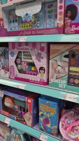 The video displays a toy store aisle filled with children's toys, primarily focused on beauty and role-playing. The shelves are stocked with various boxed items, including makeup sets, hair styling kits, dolls, and accessories. Brands like "Frozen" and "Little Darlings" are visible. The lighting is bright and appears to be artificial, typical of retail environments. The camera pans across the shelves, offering a close-up view of the toy packaging. No people are explicitly shown in the video, although the toys themselves feature images of children and dolls. The overall environment is clean and organized, suggesting a well-maintained retail space.