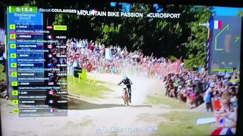 The video shows a screen displaying a live broadcast of a mountain bike race. The screen features a leaderboard on the left side, listing racers and their times, with Benoit Coulanges currently in first place. In the center of the screen, a mountain biker, wearing a black and blue helmet and jersey with red accents, is riding down a dusty, sloped track. Spectators line the sides of the track, cheering. The broadcast also shows replays and graphics, including logos for "MOUNTAIN BIKE PASSION" and "EUROSPORT," as well as national flags. The text "COULANGES FOR THE WIN" is displayed at the bottom of the screen. The footage is from a television or monitor, with some slight glare and reflections visible on the screen. The overall impression is a competitive sporting event with an excited crowd.