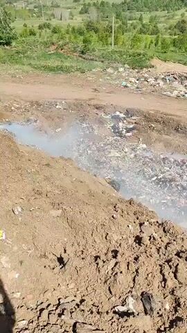 The scene presents a large, open landfill characterized by a significant accumulation of diverse waste materials. Visible in the center of the frame is a considerable pile of garbage, including plastics and other unrecognizable items. Emanating from the landfill is a plume of smoke, suggesting combustion or decomposition is occurring. The area is surrounded by mounds of earth and a dirt road, which appears to be used for access and management of the landfill. In the background, there is a landscape of green trees and vegetation. The overall environment seems rural or agricultural, with the landfill situated in a partially developed area. The video is captured from a stationary position, providing a comprehensive view of the scale and characteristics of the landfill. The weather appears to be clear and sunny.