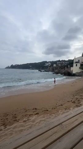 The video shows a person walking into the ocean on a sandy beach on an overcast day. The beach is framed by a wooden deck in the foreground and a rocky coastline with buildings in the background. Waves are gently lapping onto the shore. The person raises their arms briefly while standing at the water's edge.