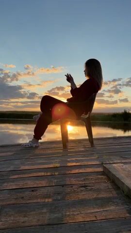 A young person sits in a chair on a wooden deck overlooking a body of water during sunset. The person is silhouetted against the setting sun, which is casting warm, golden light across the sky and reflecting on the water. The person is holding a phone and appears to be using it. The sky is filled with dramatic clouds that are illuminated by the sunset. The overall atmosphere is peaceful and serene.