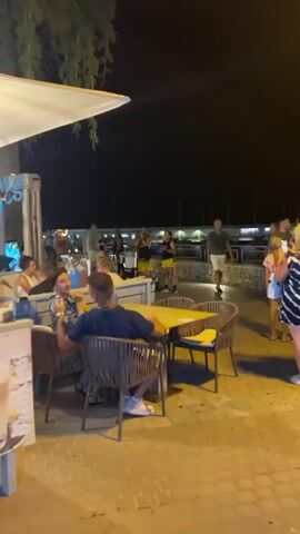 This video shows an outdoor dining area at night. In the foreground, two people are seated at a table with chairs. The table is yellow, and the chairs are woven. A person on the left is holding a blue tablet. In the background, a group of people are gathered, some standing and some seated. Some appear to be performing music. There is a palm tree visible on the left side of the frame. The ground is paved with bricks. Lights illuminate the area.