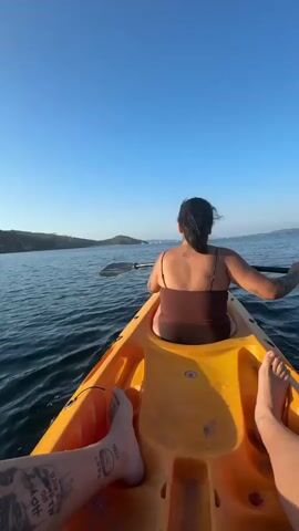 The video shows a first-person perspective from inside a yellow kayak on the water. A person wearing a brown swimsuit and with dark hair tied back is in the front of the kayak, paddling with a black oar. The water is blue and slightly rippled. In the distance, there is a rocky shoreline with some vegetation and trees. The sky is clear and blue, with the sun shining brightly and reflecting on the water. The camera is positioned low, showing the legs of another person (presumably the videographer) in the front of the kayak, with tattoos visible on one leg. The overall scene depicts a peaceful kayaking experience on a sunny day.