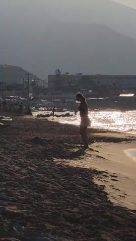 A person in a bikini performs a backflip on a sandy beach. The sun is setting, casting a warm glow over the scene. The ocean is in the background, with gentle waves lapping at the shore. There are other people in the distance, as well as some buildings and trees.