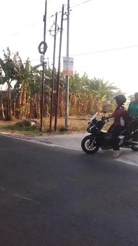 The video begins with a shot of a road with a white line, flanked by vegetation and utility poles. It then transitions to a first-person perspective from a motorcycle, showing the road ahead and passing by trees and houses. Other motorcycles are visible in the distance and passing by. The rider's hand is on the handlebars, and the motorcycle's mirror is visible. The environment appears to be a rural or suburban area with a clear sky.