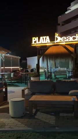 The video shows an outdoor seating area of a restaurant or bar at night. The area is illuminated by warm lighting from various sources, including string lights, spotlights, and illuminated signs. A prominent sign reads "PLATA Beach". There are multiple seating areas with wooden tables and comfortable-looking sofas and chairs. Some of the seating areas have small, lit decorative elements on the tables. In the background, there is a building with a thatched roof structure and a more modern section with windows. People can be seen inside the building and at some of the outdoor tables. The ground is paved with bricks and there is grass in the foreground and background. The overall atmosphere appears to be relaxed and inviting, suitable for evening dining or socializing.