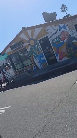 The video shows a daytime exterior shot of Martha's Corner in Hermosa Beach. A red bike is parked near the building. There is a stop sign on a post and a sign that reads "May Use Full Lane" with a bicycle symbol. Cars are parked on the street. Green umbrellas cover outdoor seating. The building has a mural with images of the sun, palm trees, and marine life.