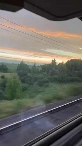 A video captures a view from a moving train. The camera looks out of a window, showing the landscape passing by. The sky is filled with soft, wispy clouds in hues of orange, pink, and gray, indicating either sunrise or sunset. Below the sky, a line of trees and vegetation creates a green border. In the foreground, train tracks are visible, reflecting the light. The motion of the train causes the trees and landscape to appear blurred. The perspective is from inside the train compartment, with the window frame visible at the top and bottom of the screen.