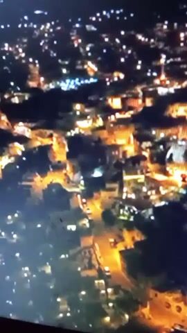 The video provides an aerial view of a town at night. The scene is dominated by the lights of buildings and streetlights, creating a warm, orange glow against the dark sky. There are hints of roads and vehicles, suggesting the town is active even after dark. The overall impression is one of a bustling community illuminated by artificial light.