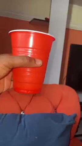 A hand holds a red plastic cup with a white rim in front of an orange tufted chair. A blue pillow is partially visible at the base of the chair. The background includes a peach-colored wall, a tall white bookshelf with multiple shelves, and a television.