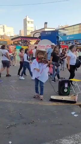 A young child dressed in a cowboy hat and a white poncho with red trim stands in a paved outdoor area, playing a violin. A black portable speaker and an open violin case are on the ground in front of the child. Several adults and children are walking in the background, some looking at the child. Buildings with storefronts are visible behind the people. The sky is clear and the sun is shining. The overall scene appears to be a public plaza or street market.