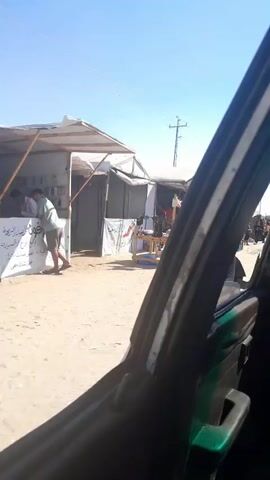 The video shows an outdoor market scene. Several makeshift stalls with canvas roofs are set up along a dirt road. Goods are displayed on tables and on the ground. There are people walking and standing around the stalls. In the foreground, part of a vehicle is visible, suggesting the footage is taken from inside a car. A utility pole with wires is visible in the background against a clear blue sky.