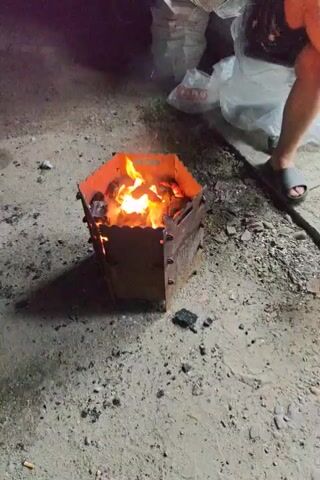 A hexagon-shaped metal brazier sits on the ground, filled with burning coals and emitting sparks. A person's hand holding a pair of pliers is visible in the foreground, with the pliers positioned near the brazier, suggesting they are interacting with the coals. The ground appears to be a mix of dirt and gravel. In the background, there are some white bags and a person wearing sandals. The scene is dimly lit, indicating it is likely evening or nighttime.