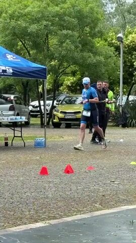 Here is a description of the video:

The video shows a man running in what appears to be a triathlon. He is wearing a blue and black tri suit, a white cap, and running shoes. He is running on a gravel surface marked with red and yellow traffic cones. 

To his left is a table with water bottles and other drinks. Behind the table is a person in a black jacket pouring a drink from a bottle. Above the table is a blue tent with "TKPS Triathlon Klub Poznanica" printed on it. Behind the tent are parked cars and trees. 

In the background, there is a tall stone structure that looks like a tower or part of a castle. 

The scene is outdoors, and the weather looks overcast. There is another person walking with an umbrella to the right of the runner.