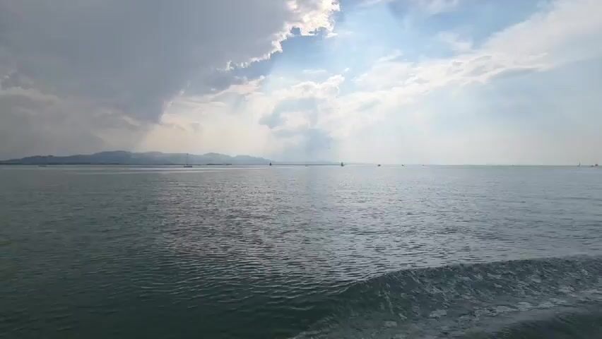 The video shows a view from a boat moving across a body of water. The water is choppy with a wake visible on the right side of the frame. In the distance, a landmass with mountains can be seen. The sky is partly cloudy with some rays of sunlight breaking through. There are a few sailboats visible on the water in the distance. The overall impression is one of a vast, open body of water under a dynamic sky.