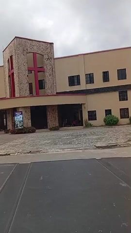 The video shows the exterior of a modern church building. The building is made of beige-colored stucco and features a prominent stone-clad tower with a large red cross. The entrance area is covered by a canopy. In front of the building, there is a paved courtyard with some people walking around. To the left of the building, there is a large billboard with the word "ANAZOSIS" in blue letters. Several people are visible in the video, including individuals standing near the entrance and others walking in the courtyard. The sky is overcast, suggesting a cloudy day. The camera appears to be positioned from a vehicle, as the bottom of the frame shows a dark surface, possibly a car's dashboard or hood.