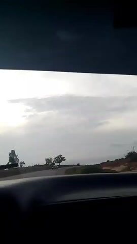 The video captures a view from inside a vehicle looking out the windshield. The scene is a rural landscape under a cloudy sky. A car is visible on a curved road in the distance. Several trees dot the landscape. A car air freshener hangs from the rearview mirror, partially obscuring the view. The video appears to be recorded while the vehicle is in motion.