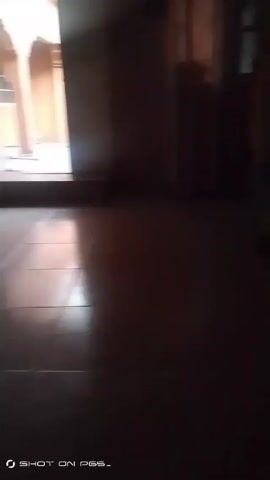 The video shows a dark interior space with a blurry view towards a lighter area. There appears to be a hallway or opening leading to a brightly lit outdoor area. There are glimpses of walls, flooring, and possibly furniture or construction materials. The camera moves slightly, showing more of the dark interior and then focuses on a dark surface, possibly fabric or a wall. Someone says "Amen" repetitively in the background.
